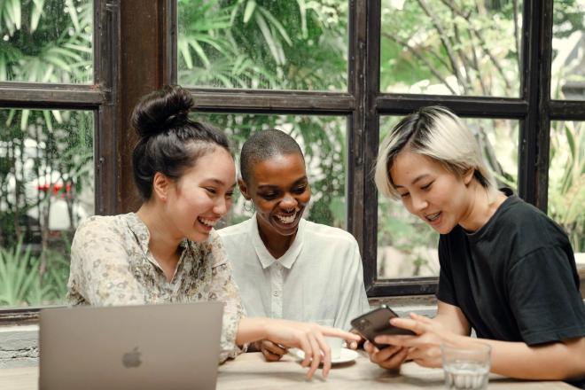 Drie mensen zitten rond een tafel in een café achter een laptop, samen lachend, en wijzen naar een mobiele telefoon. Afbeelding door Ketut Subiyanto op Pexels.
